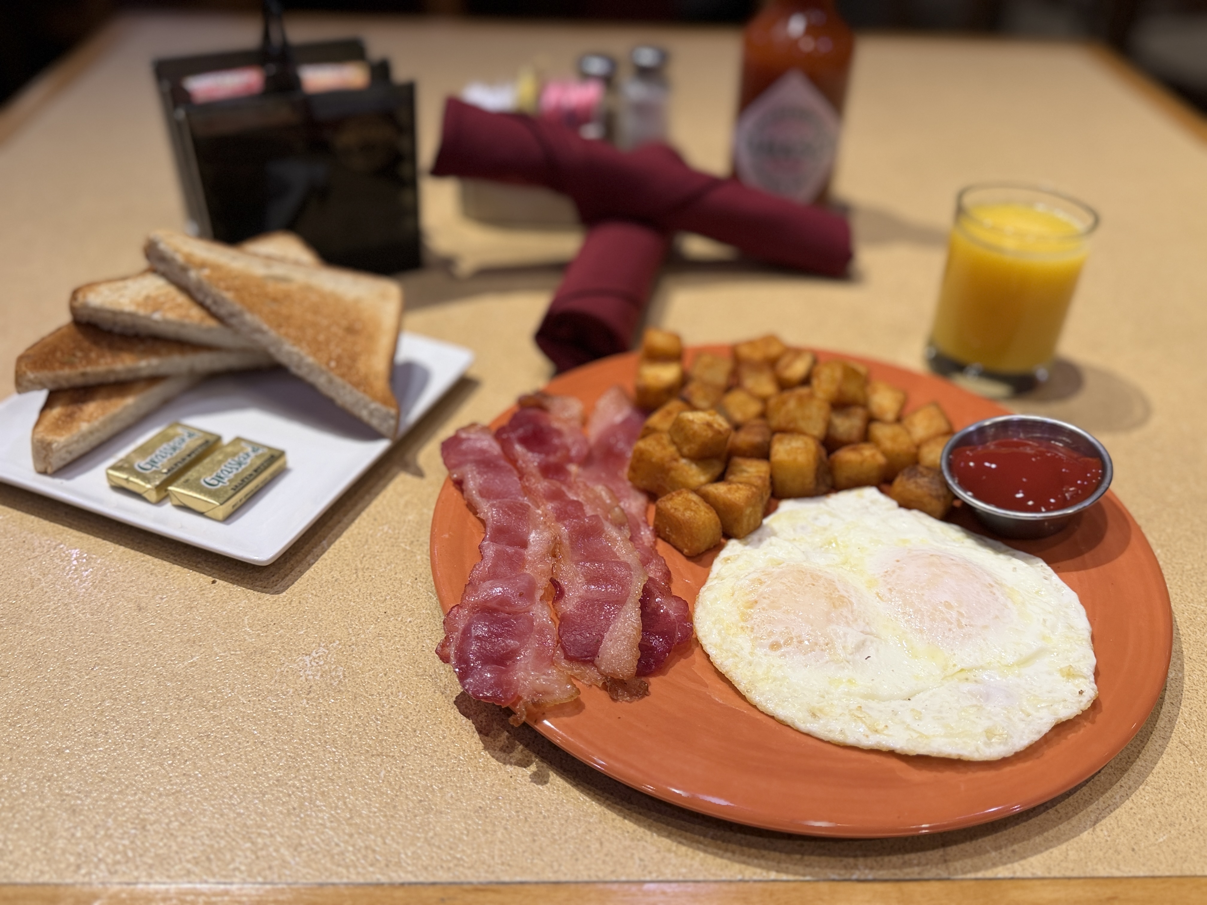 Image of breakfast plate with eggs, bacon and home fried potatoes. Glass or orange juice. Side plate of toast with butter