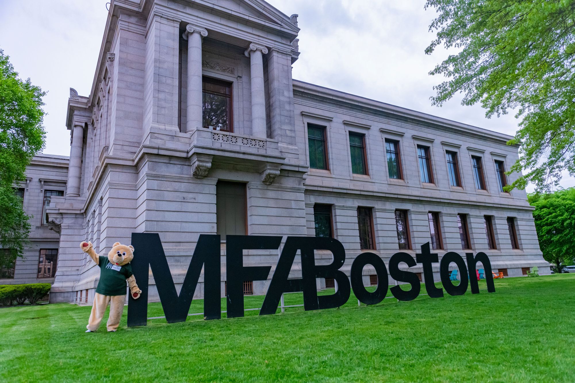 A large stone building with a sign reading MFA Boston in front of it, and a mascot bear standing next to the sign.