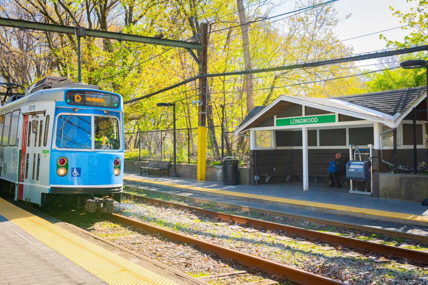 A blue and white train stopped at a station with a green sign reading 'Longwood'.
